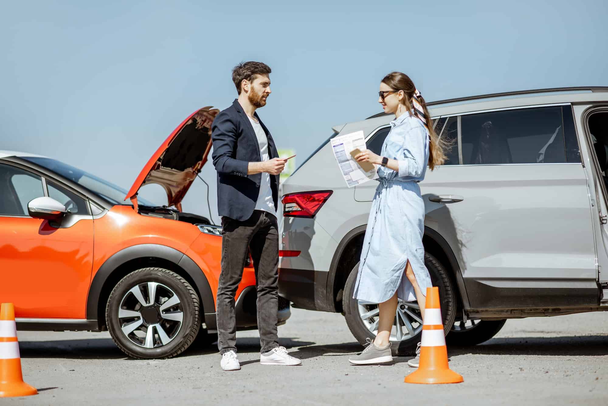 A man and woman standing next to an orange car with its hood open.